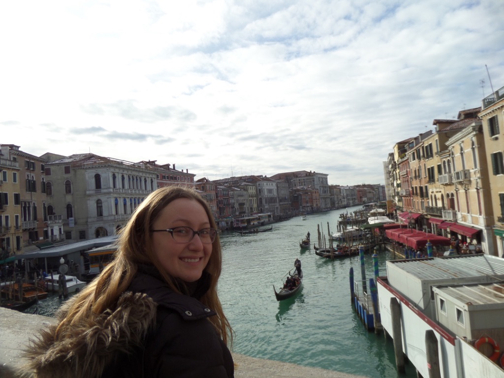 Person standing on the Rialto Bridge in Venice, Italy, with the Grand Canal and historic Venetian buildings in the background.