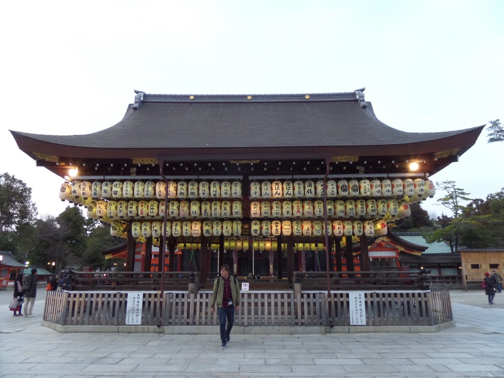 Man standing in front of the Yasaka Shrine in Kyoto, Japan, with its iconic red torii gate and traditional architecture.