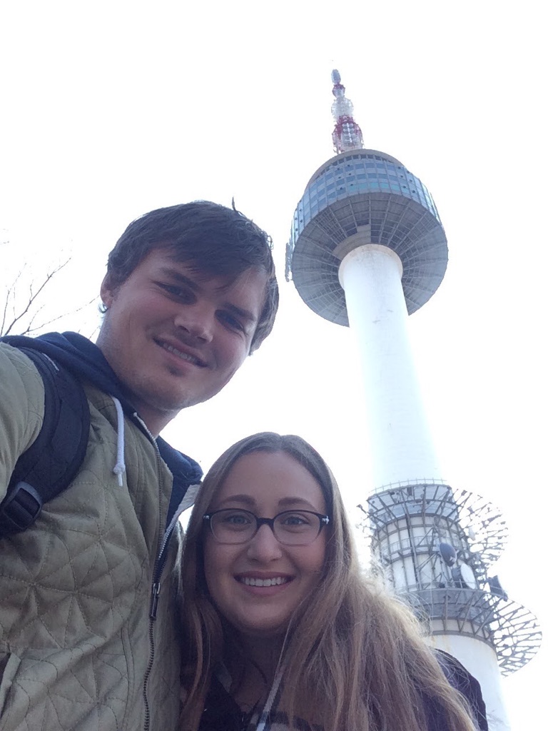 Couple taking a selfie with Seoul Tower visible in the background on a clear day.